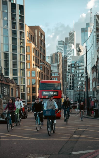 A group of cyclists riding on a city street with modern high-rise buildings on both sides, including glass-fronted towers and brick structures, under a partly cloudy sky. In the background, a red double-decker bus displaying route number 26 towards Hackney Wick is approaching. The scene features pedestrians and vehicles, with some cyclists wearing casual clothing and helmets, and others carrying bags. The urban environment reflects a busy, contemporary setting typical for house removals and furniture transport in Hackney Marshes, with the area suitable for logistics involved in home relocation services offered by Man with Van Hackney Marshes.
