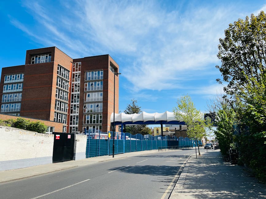 Photograph of a multi-storey brick residential building viewed from the street on a clear day with blue sky and scattered clouds. In the foreground, a paved road runs alongside a white brick wall with a black metal gate. To the right, there is a row of small trees with light green leaves, casting shadows on the pavement. A covered bus stop with a wave-shaped white roof and supporting poles is located adjacent to the building, and a blue construction fence runs along the sidewalk. The scene captures an urban environment, potentially during a home relocation or furniture transport process, with no vehicles or moving equipment visible, highlighting the surrounding infrastructure and urban landscaping. The image is associated with house removals and moving services, typical of a professional relocation setting.